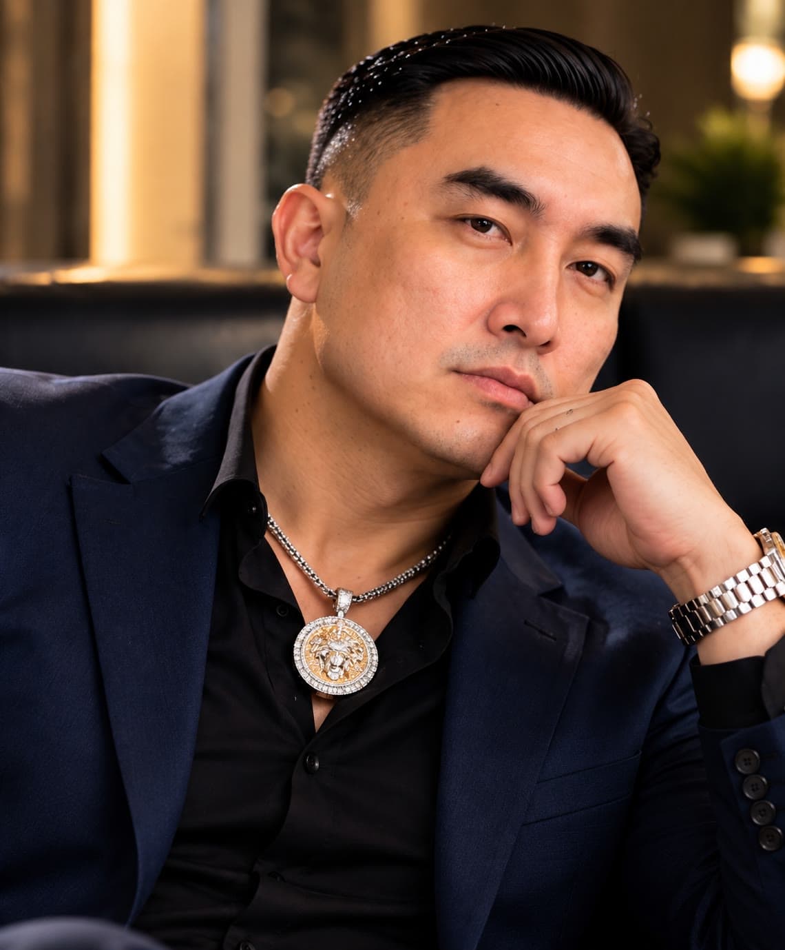A man in a navy suit and black shirt wearing a silver chain with a gold medallion pendant and a silver link watch