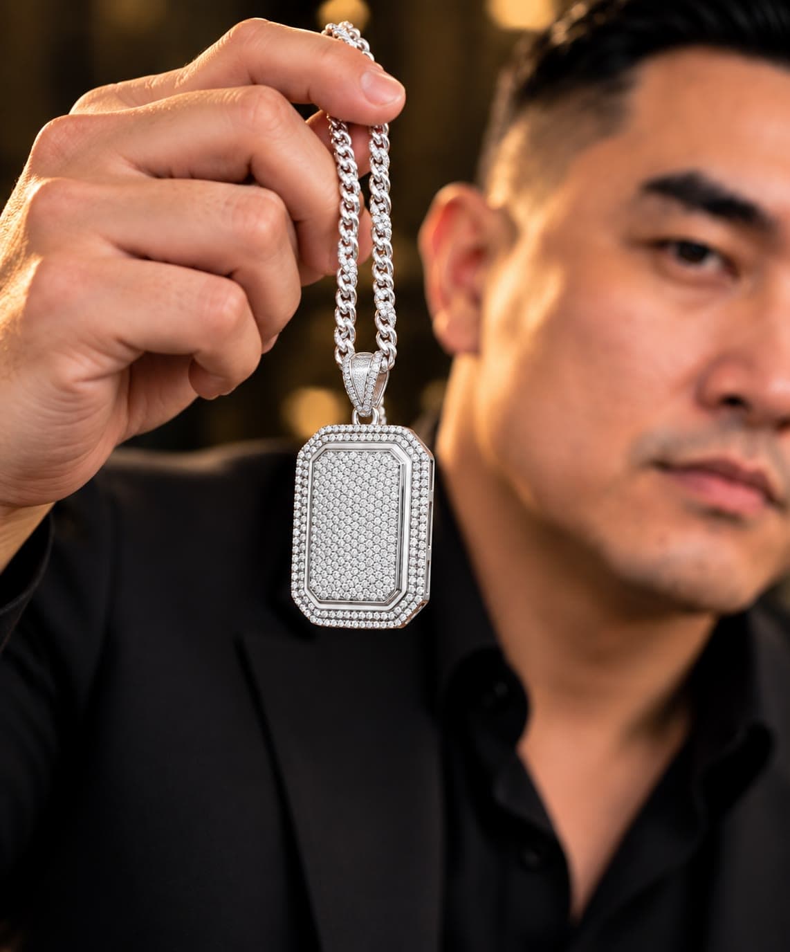 A man in a black suit holding a silver Cuban chain with a large iced rectangular pendant